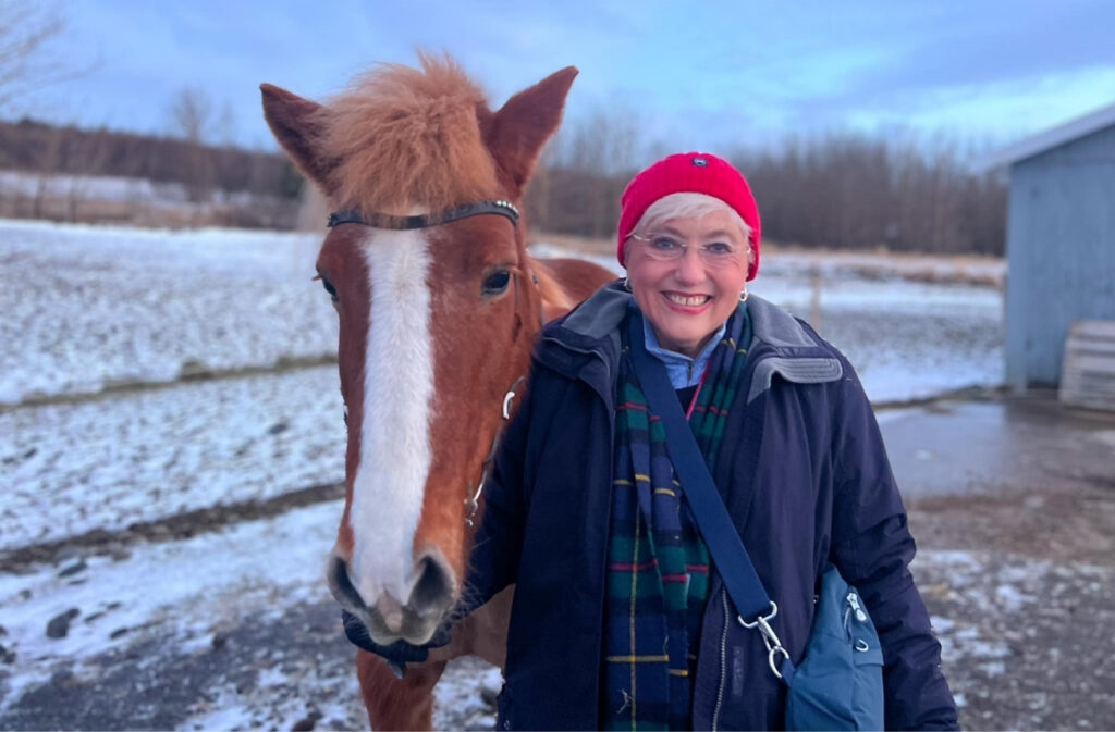 Older woman in a red beanie posing with a chestnut pony in a snowy farm setting, smiling at the camera.