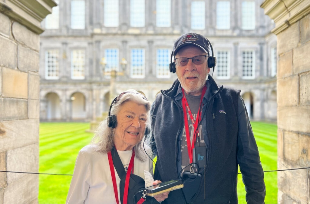 Two smiling seniors wearing headphones stand in a stone archway courtyard with a green lawn behind them, posing for a photo with lanyards.