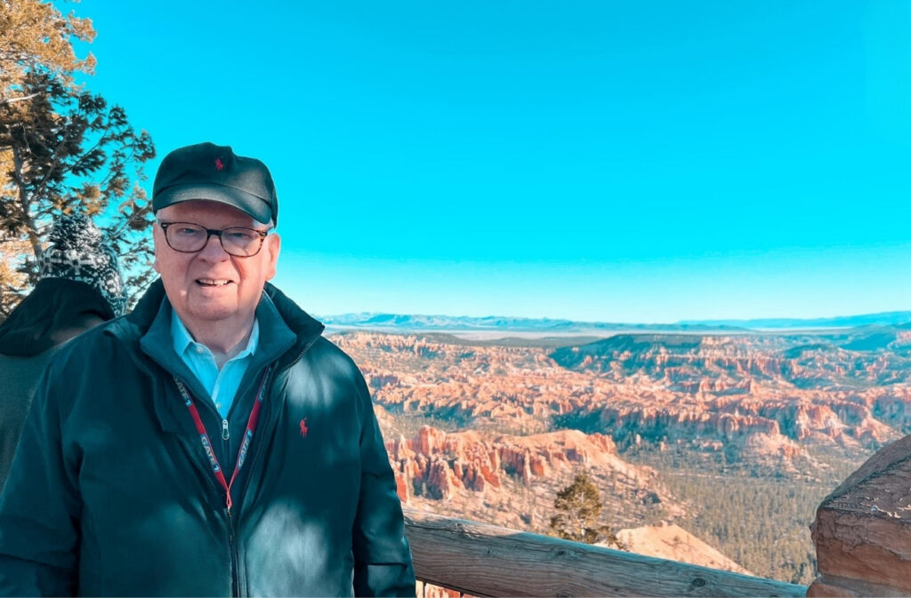 Older man wearing a dark jacket and cap stands at a canyon overlook with red rock formations and a bright blue sky behind him.