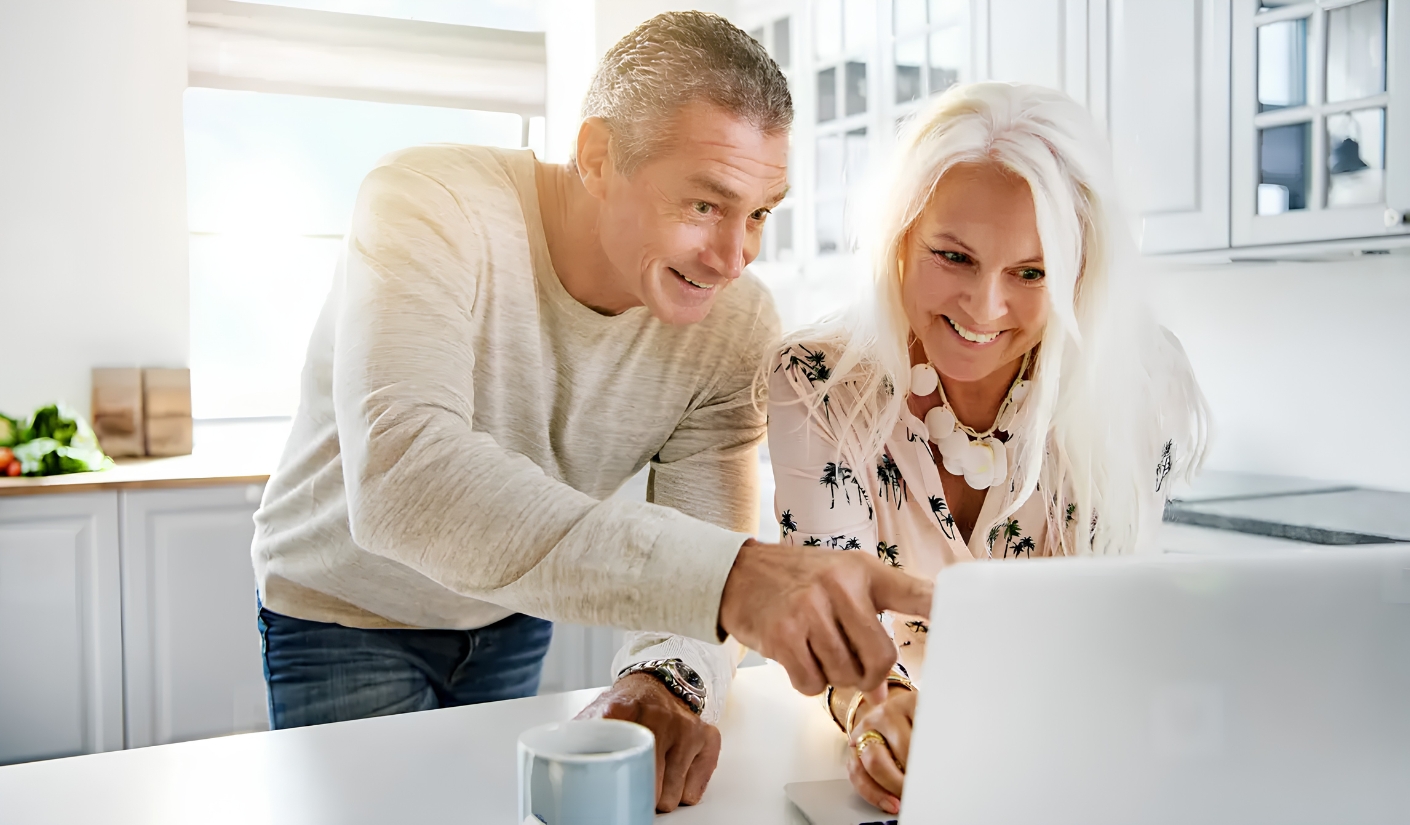 Smiling couple standing in a bright kitchen, pointing at a laptop on the counter.
