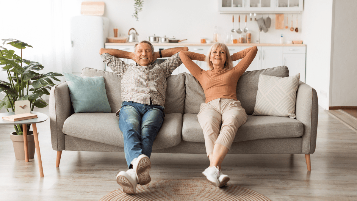 Older couple lounging on a gray sofa with arms behind heads, smiling in a bright, cozy living room with a plant nearby and a kitchen in the background.