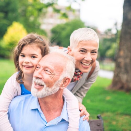 Grandfather with a granddaughter on his shoulders, smiling in a sunny park with grandma beside them on a bench in the background.