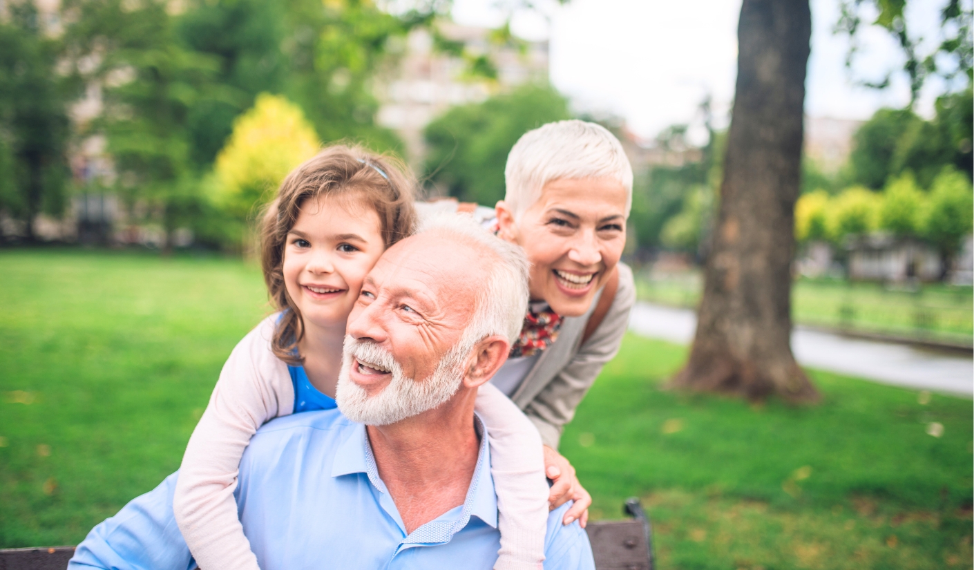 Grandfather with a granddaughter on his shoulders, smiling in a sunny park with grandma beside them on a bench in the background.